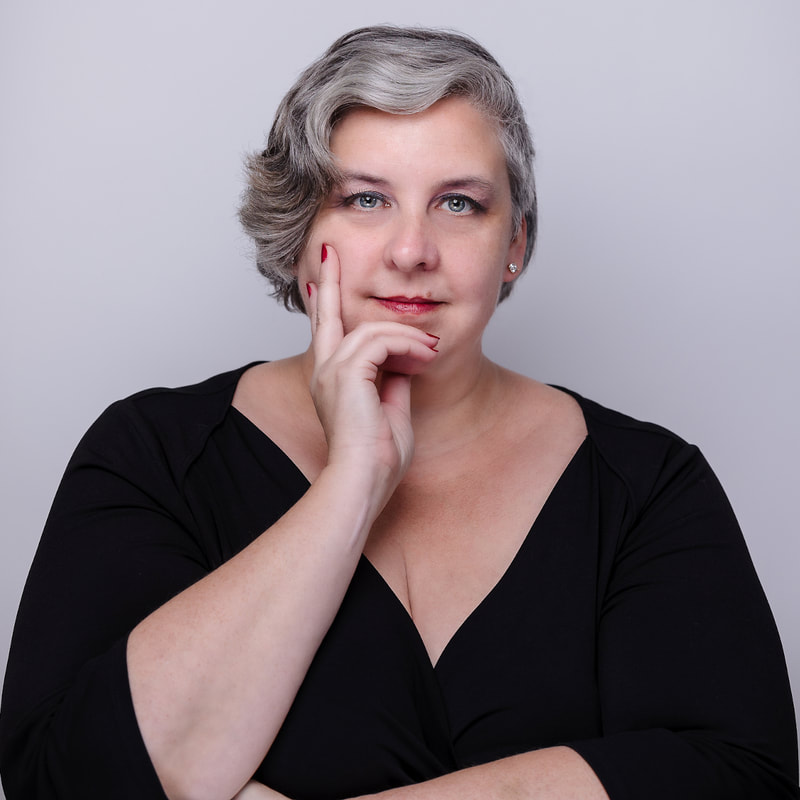 Photo of Jessica Ellis-Wilson, a pluz-sized white nonbinary person with blue eyes and short grey hair swept to the left, looking directly at the camera and resting their chin on their right hand while their left arm rests across their body with their left hand under their right elbow.  They are standing in front of a grey-white background in a black wrap shirt with 3/4 sleeves. They have red nail polish on short nails, matching red lipstick, and a single stud earring is visible in their left ear.