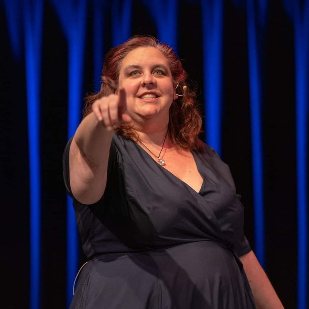 Photo of speaker Jessica Ellis-Wilson: a plus-sized white nonbinary person with blue eyes and med-length curly brown hair swept back from their face, looking above and to the left of the camera and smiling. They are standing in front of a blue stage curtain pointing toward the camera with their right hand. They are wearing a navy blue wrap dress with a pendant on a black cord around their neck. A headset microphone is visible around their left ear.
